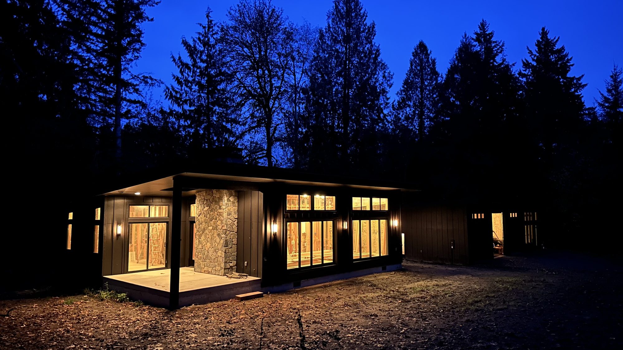 The house build at dusk, warm light through black-framed windows against the forest